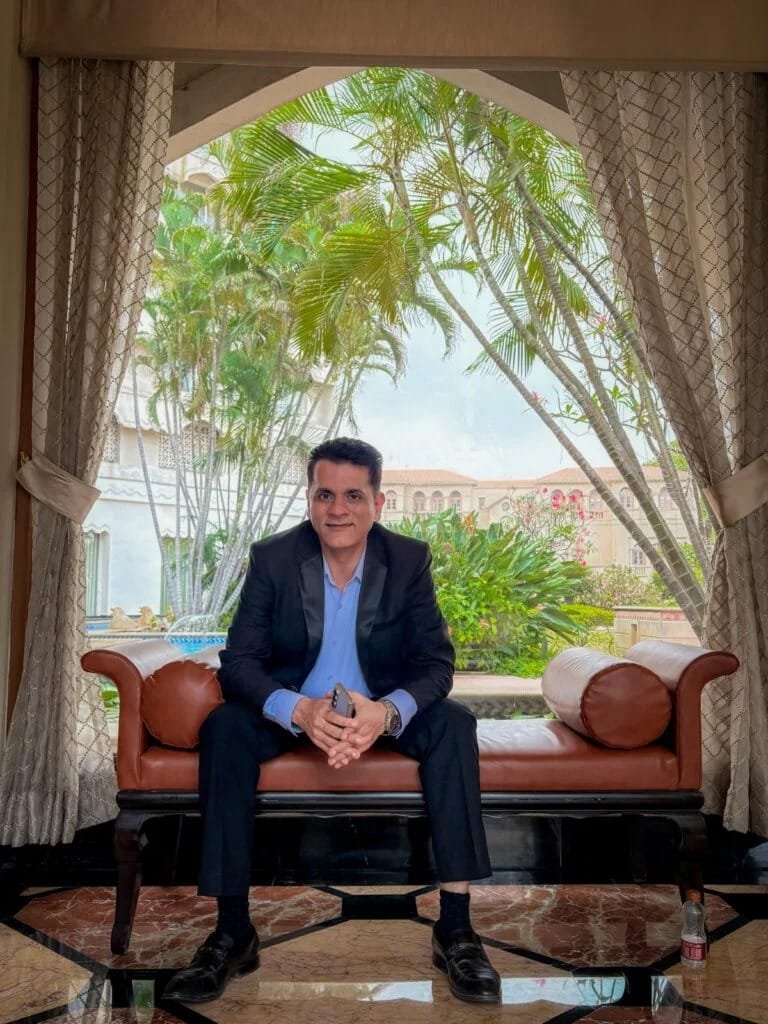 Professional businessman in navy suit seated on leather couch in luxury hotel lobby with palm trees and arched windows