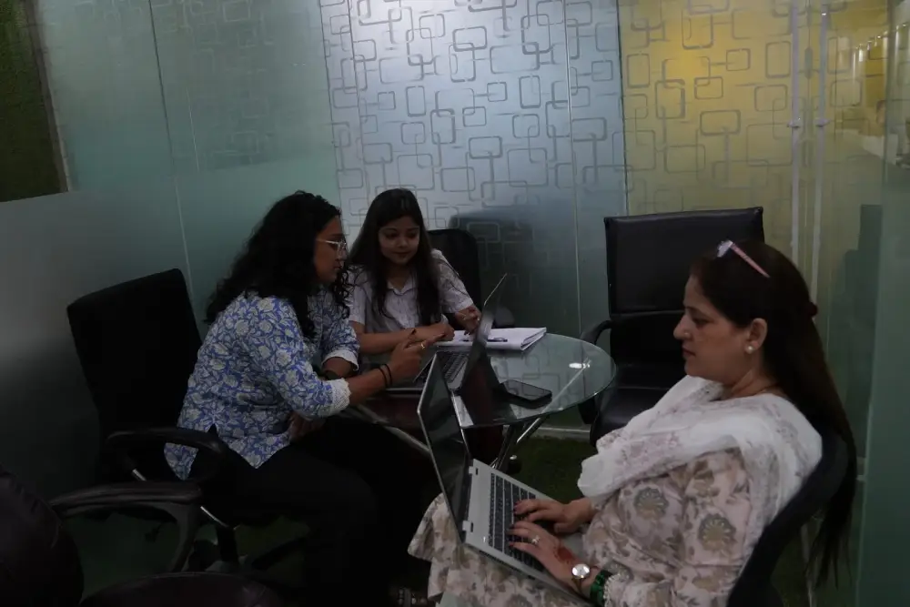 Three professional women collaborating on laptops in a modern office meeting room with geometric wall design