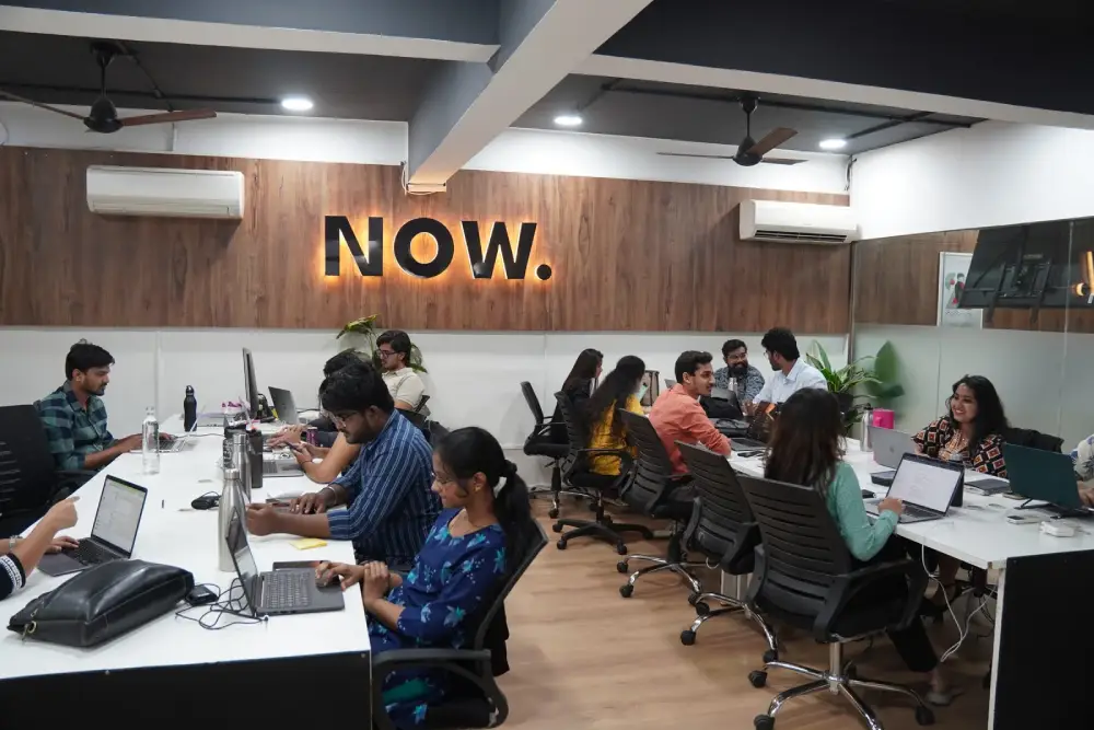 Modern office workspace with collaborative team working on laptops and computers at desks with NOW sign